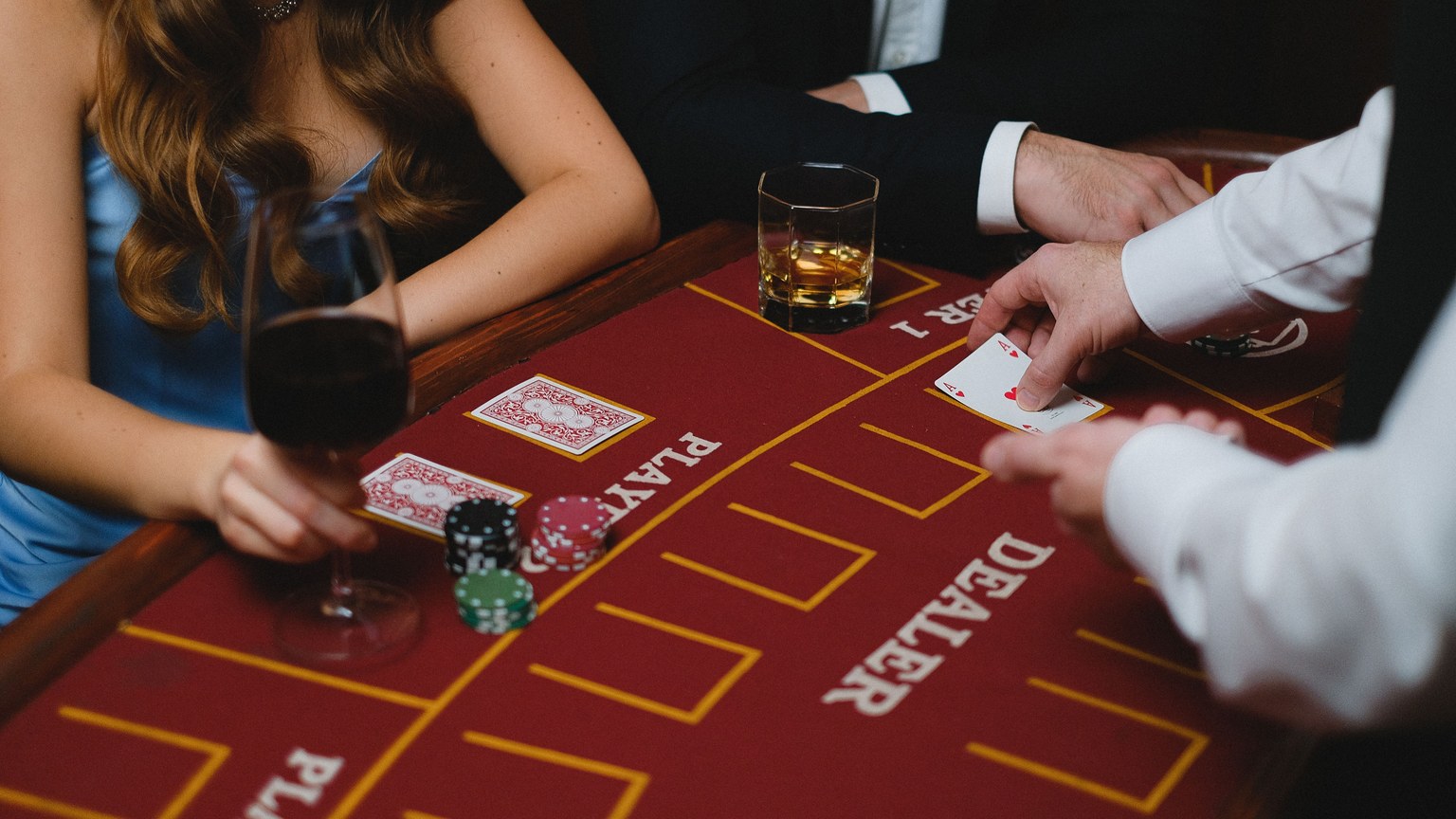 Close-up of a card game with players and chips at a casino table, featuring wine and whiskey glasses.