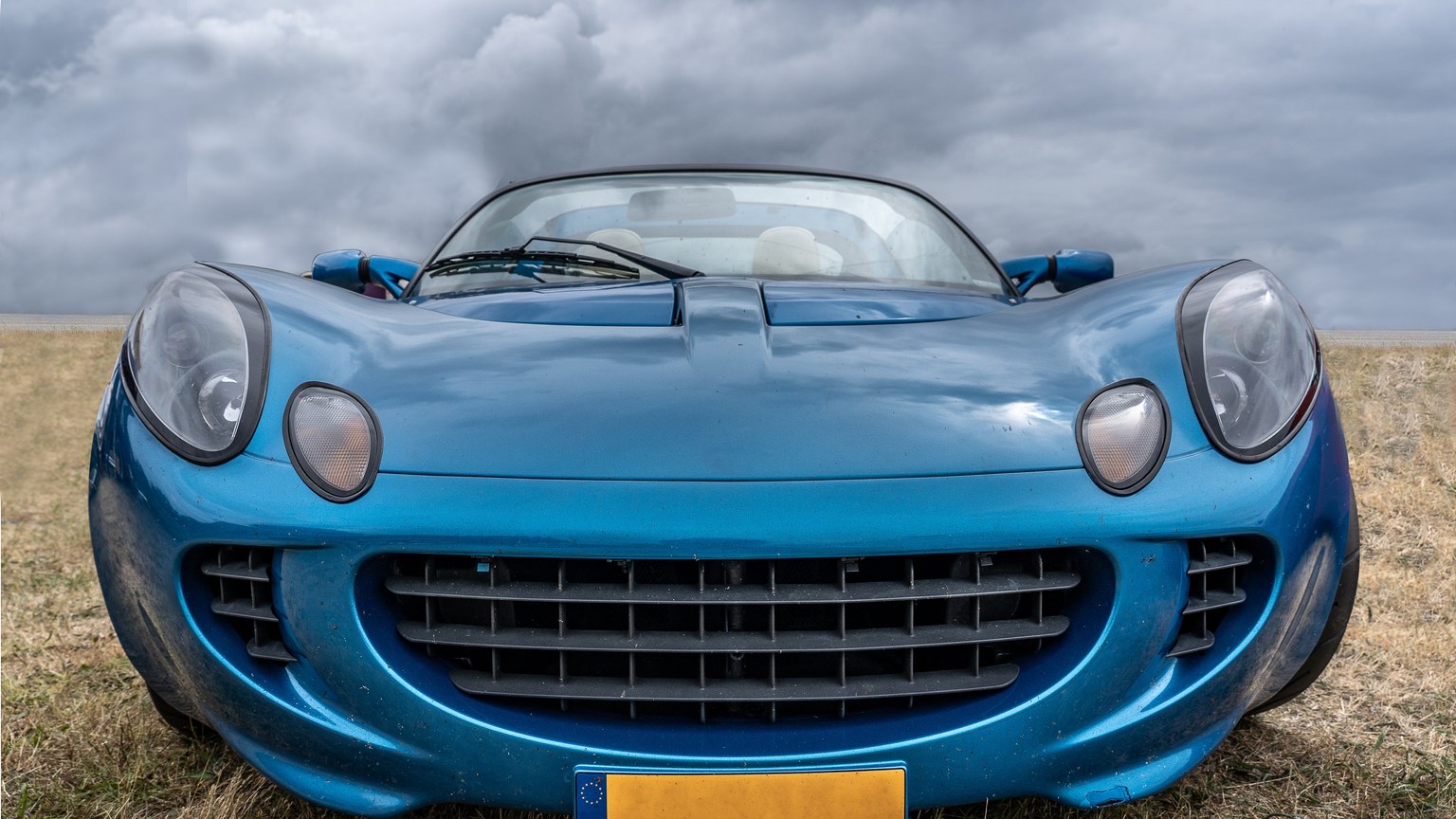 Front view of a classic blue sports car in an open field under a dramatic cloudy sky.