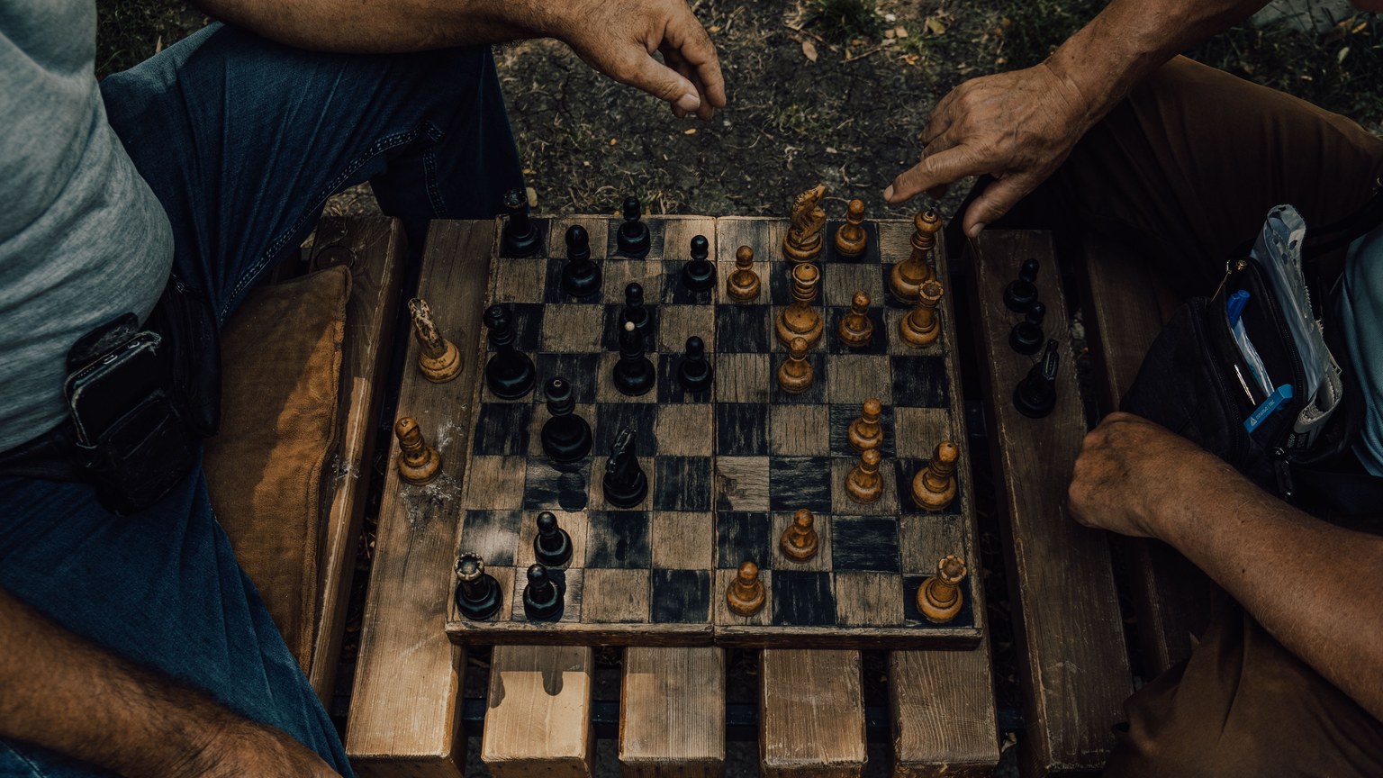 Two adults engaged in a strategic chess match on a rustic wooden board outdoors.